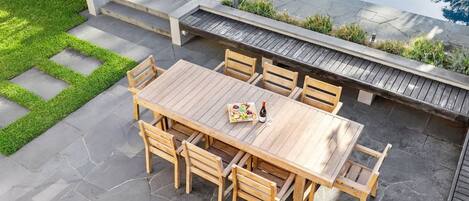 A wooden outdoor dining area with a table and chairs, surrounded by a stone patio and a small grassy area.