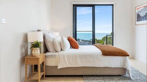 Ein Schlafzimmer mit Bett, Nachttisch, Lampe und Fenster mit Meerblick.