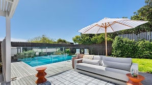 Ein moderner Poolbereich im Freien mit einer Holzterrasse, einer weißen Couch, einem orangefarbenen Tisch und einem großen Sonnenschirm.
