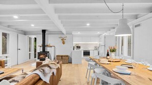 A modern kitchen with white cabinetry, a central island, and a dining area with white chairs and a wooden table. - The Coast Country Cottage (Balnarring Beach)