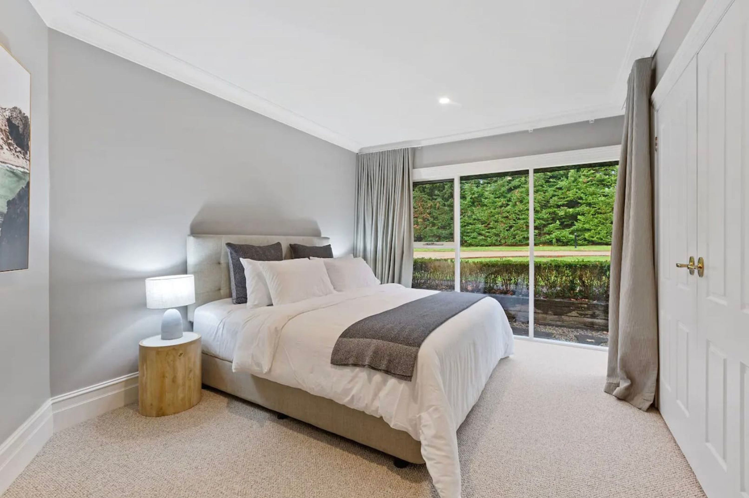 A bedroom with a large bed, a nightstand, a window with curtains, and a view of greenery outside.