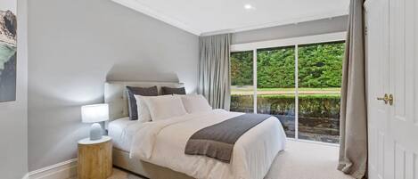 A bedroom with a large bed, a nightstand, a window with curtains, and a view of greenery outside.