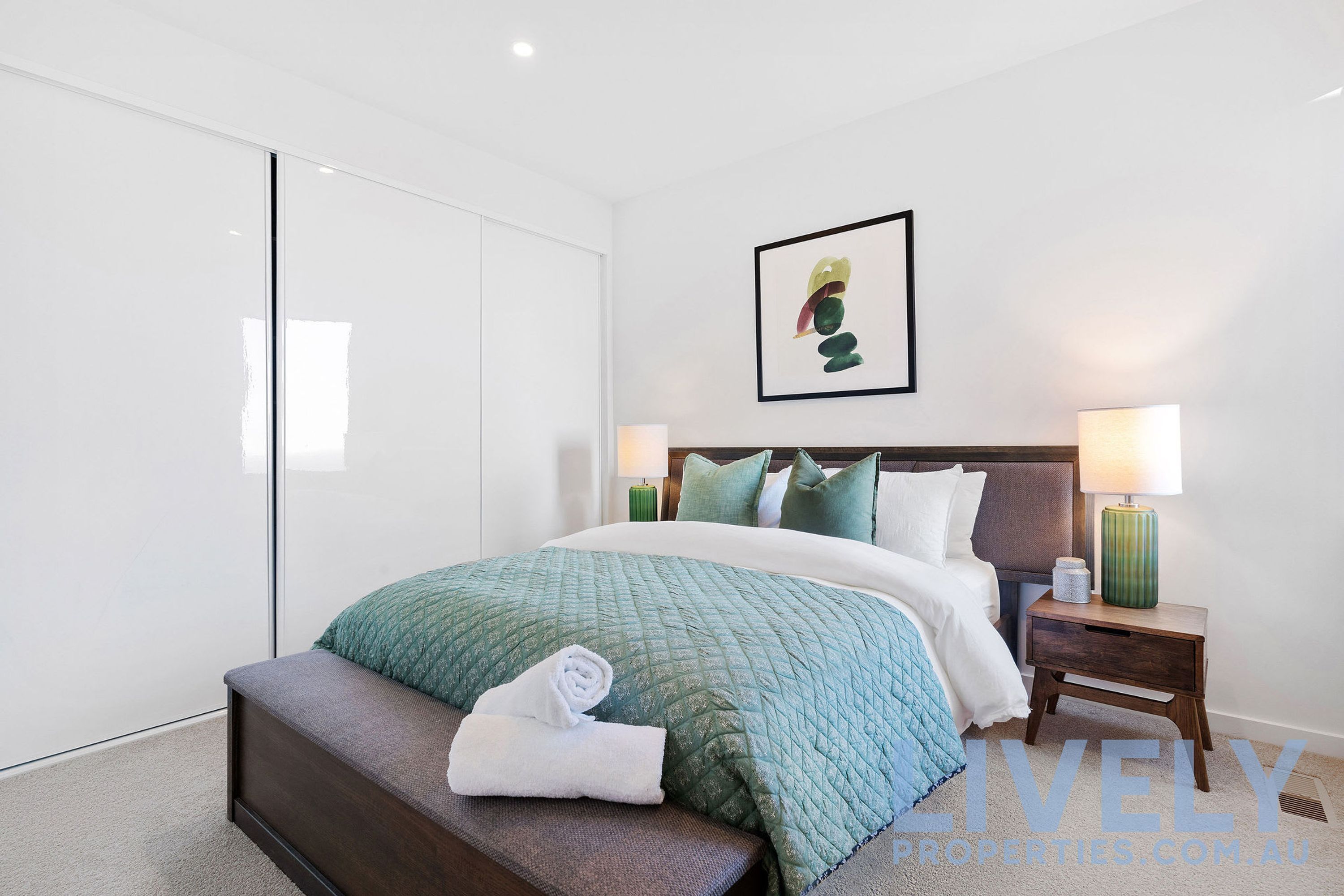 A modern bedroom with a large bed, a bench, a nightstand, and a wall-mounted artwork.