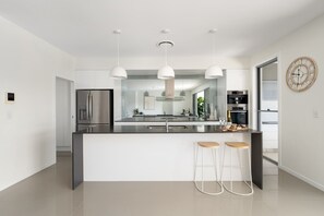 A modern kitchen with a central island, pendant lights, stainless steel appliances, and a large clock on the wall. - Luxe Family Entertainer on Canal Private Pool (Gold Coast)