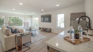 Una sala de estar moderna con un sofá, una mesa de centro y un televisor montado en la pared.  Se observa una zona de cocina con un refrigerador y un lavabo.