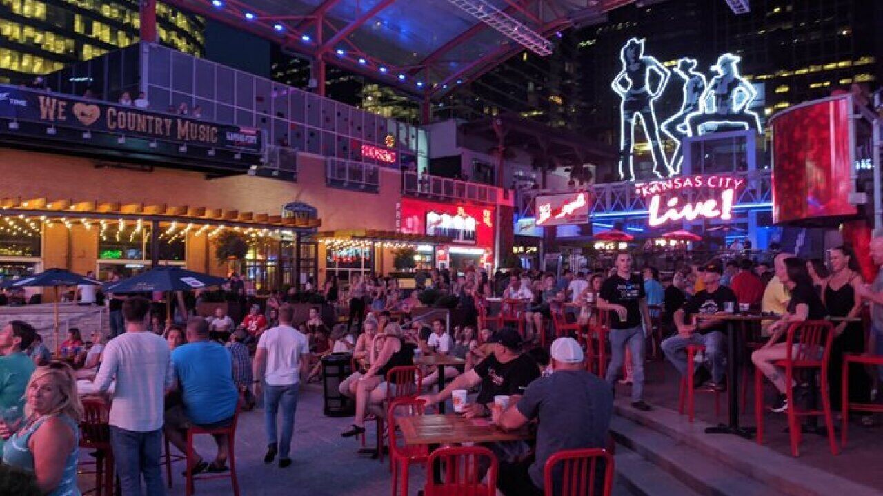 A lively outdoor dining area with red chairs and tables, surrounded by various restaurants and a large illuminated sign.