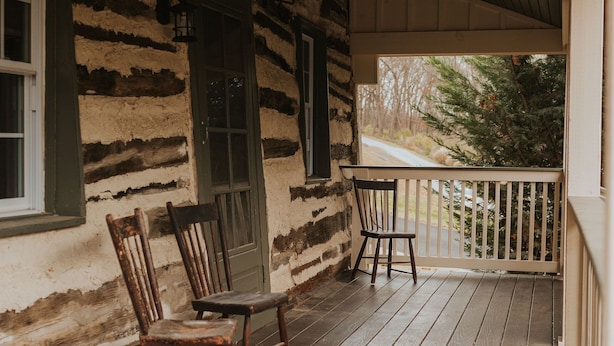 A rustic wooden porch with two wooden chairs and a small table, overlooking a road with trees and a building in the background.