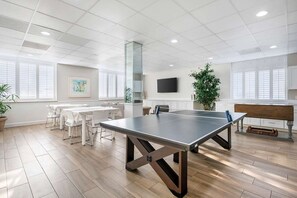 Ein geräumiges Zimmer mit einem Tischtennistisch, weißen Stühlen, einem großen Fenster und einer Topfpflanze.