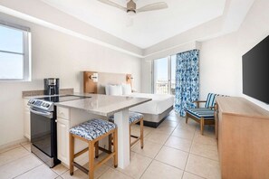 Habitación de hotel compacta con kitchenette, una cama con cabecera, zona de comedor con mesa y sillas, y un ventanal grande con cortinas.
