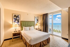 A hotel room with a large bed, two bedside tables with lamps, a view of the ocean through a balcony door, and artwork on the walls.
