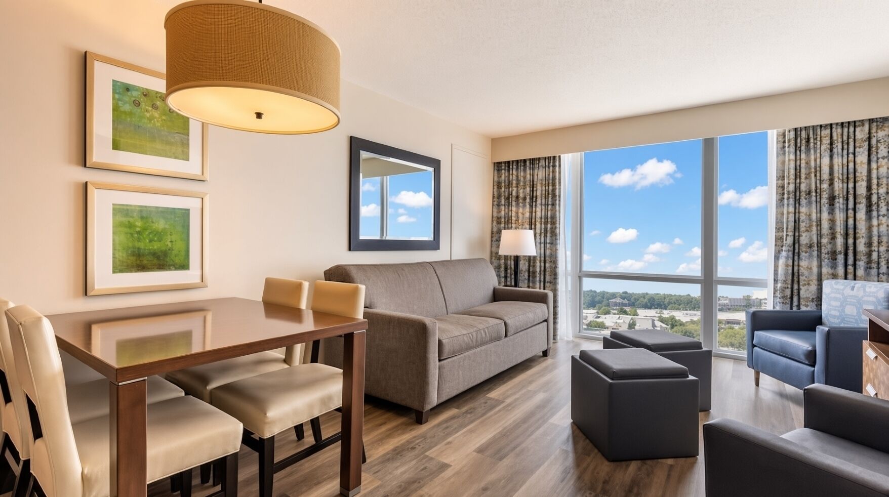 A modern hotel room with a dining area, a sofa, a large window with a view, and a hanging lamp.