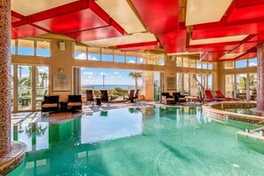 An indoor swimming pool with clear blue water, surrounded by lounge chairs and a view of the ocean.
