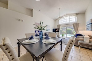 Uma sala de jantar com uma mesa de madeira escura posta para quatro pessoas, cercada por cadeiras bege estofadas. Um vaso azul com flores brancas está sobre a mesa.