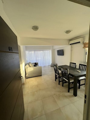 Una sala de estar con sofá, una mesa de comedor con sillas, un televisor montado en la pared y un equipo de aire acondicionado.