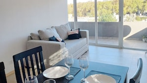 Una sala de estar moderna con un sofá, una mesa de comedor dispuesta con platos y copas de vino, y un balcón con vistas a árboles y edificios.