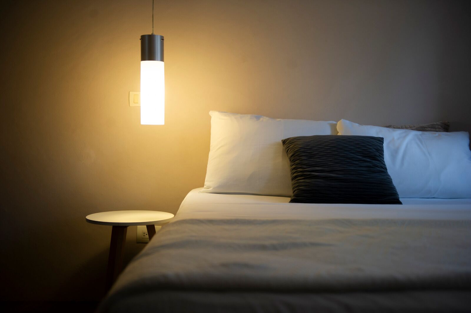 A neatly made bed with white and dark pillows, a round bedside table, and a hanging light fixture.