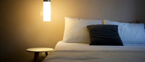 A neatly made bed with white and dark pillows, a round bedside table, and a hanging light fixture.