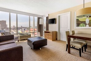 A hotel room with a large window, a flat-screen TV, a dining table with chairs, a sofa, an ottoman, and a wooden cabinet.