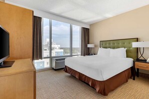 A hotel room with a large bed, a television, a desk, and a view of the city through large windows. - Diamond Resorts Ocean Beach Club - 1 Bedroom Accessible Roll-in Shower City View (Virginia Beach)