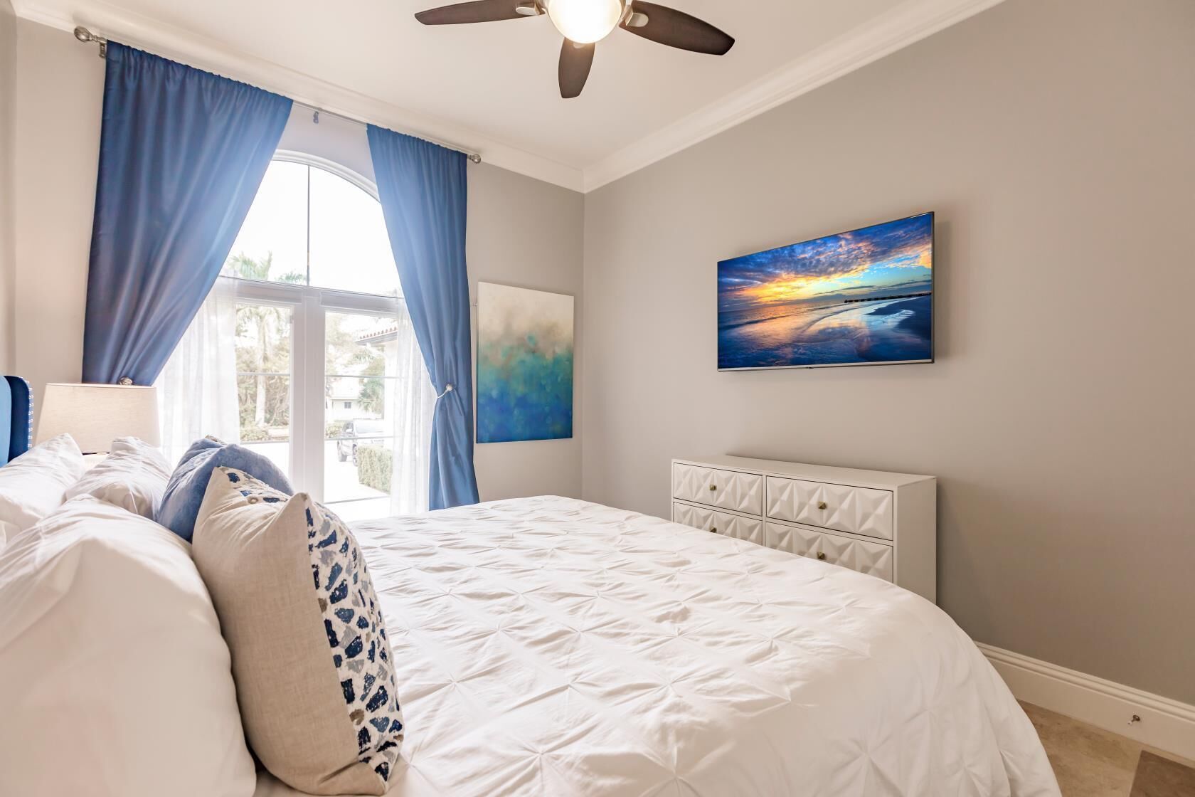 A bedroom with a bed, a ceiling fan, a window with curtains, and a painting on the wall.