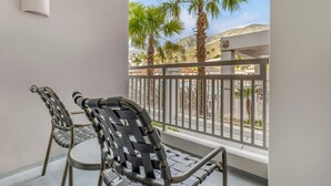 A balcony with two chairs, a white wall, and a view of palm trees and a street.