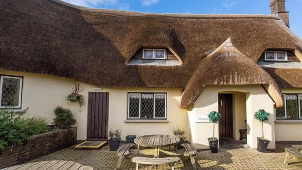 A thatched-roof cottage with a paved courtyard, a wooden bench, and potted plants. - Devon | Peaceful Country Escape | Pool + Sauna (Devon)