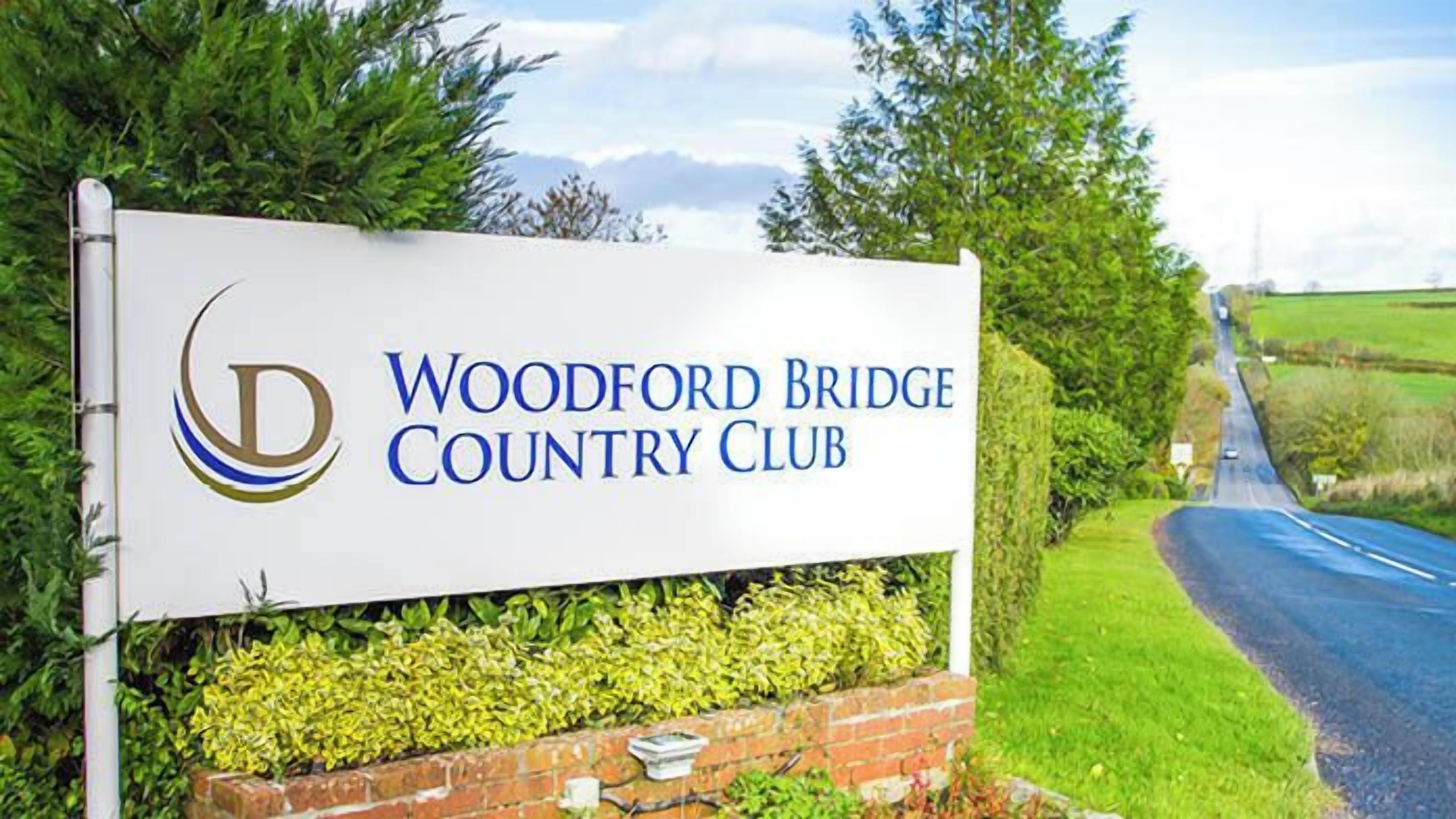 Letrero del Woodford Bridge Country Club con un logotipo, rodeado de follaje y una carretera al fondo.