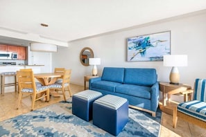 Un salon moderne avec un canapé bleu, une table à manger en bois et des chaises, une cuisine en arrière-plan et un tableau accroché au mur.