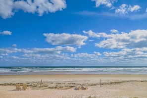 Ein Strand mit blauem Himmel, Wellen und zwei leeren Liegestühlen.