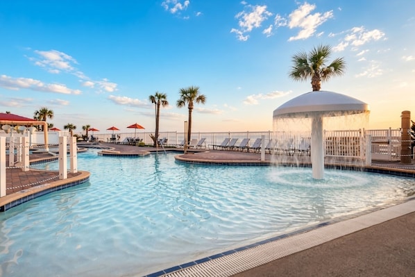 A pool area with multiple water features, including a fountain, surrounded by lounge chairs and palm trees.