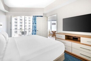 Et hotelværelse med en stor seng, et skrivebord med et fladskærms-tv, en stol og udsigt til en bygning gennem et vindue.