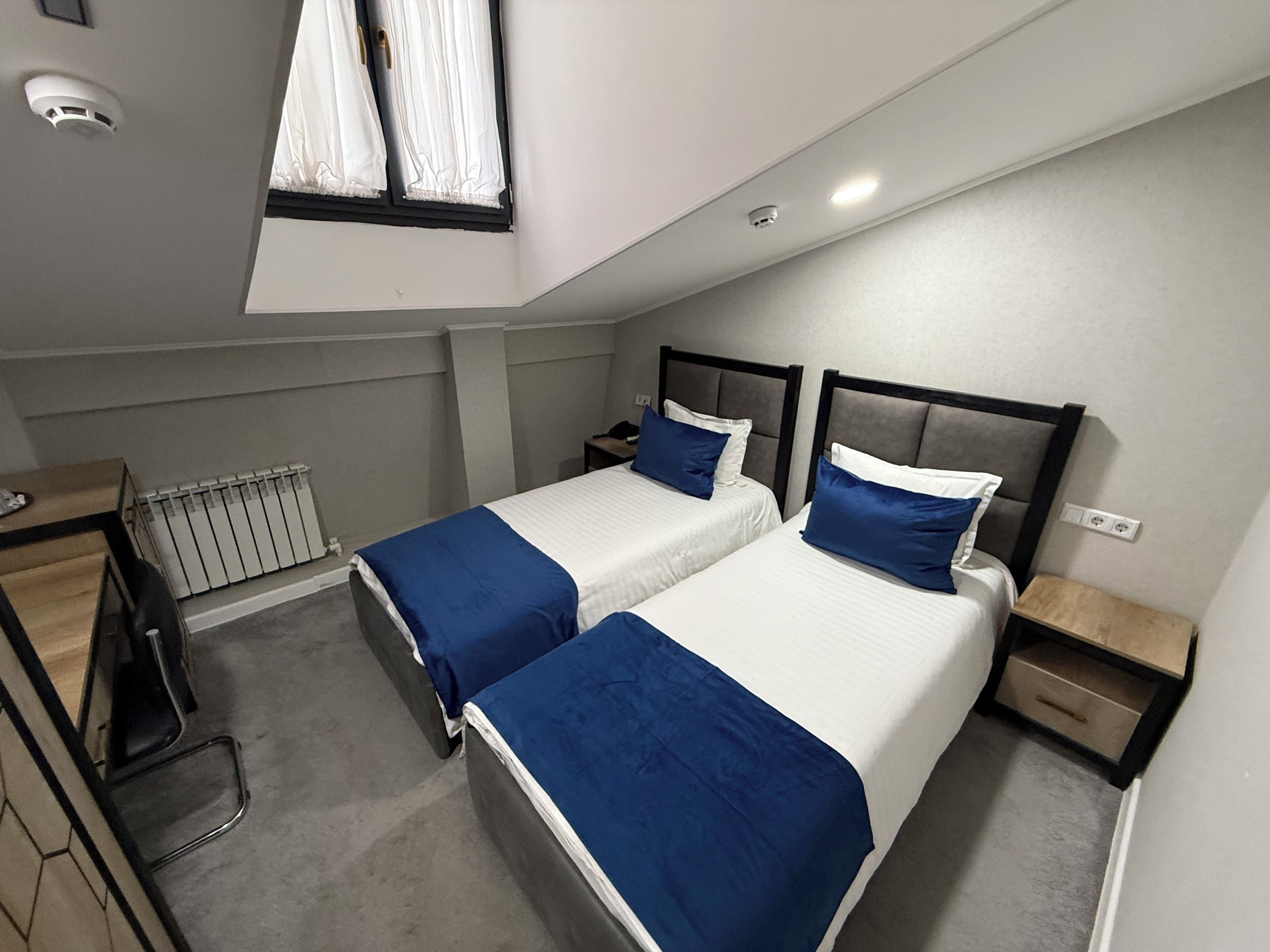 A small, modern hotel room with two beds, a desk, and a radiator.