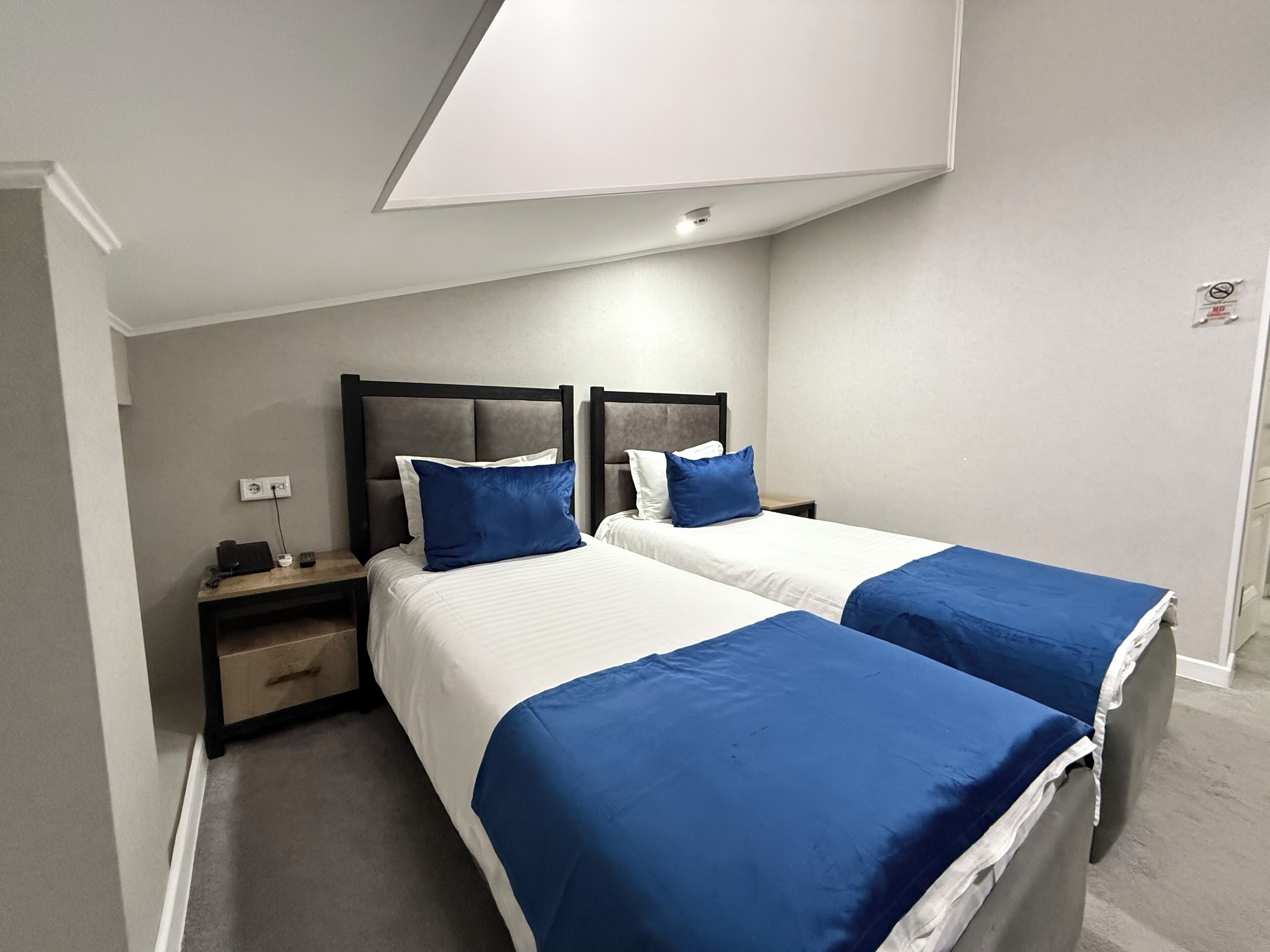 A hotel room with two beds, each with a blue bedspread and white linens. There is a nightstand with a lamp and a power outlet on the wall.