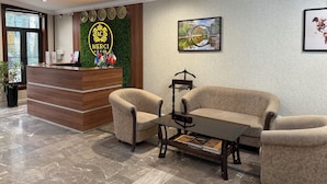 A hotel reception area with a wooden reception desk, two armchairs, a coffee table, and a framed picture on the wall.