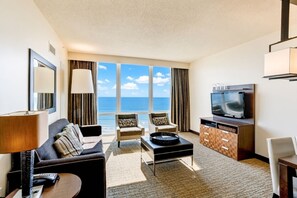 Habitación de hotel con sofá, sillones, mesa de centro, televisor y vista al océano.