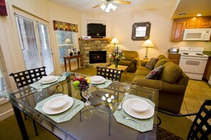 A dining area with a glass table set for four, a fireplace, a television, and a kitchen with a microwave and stove.