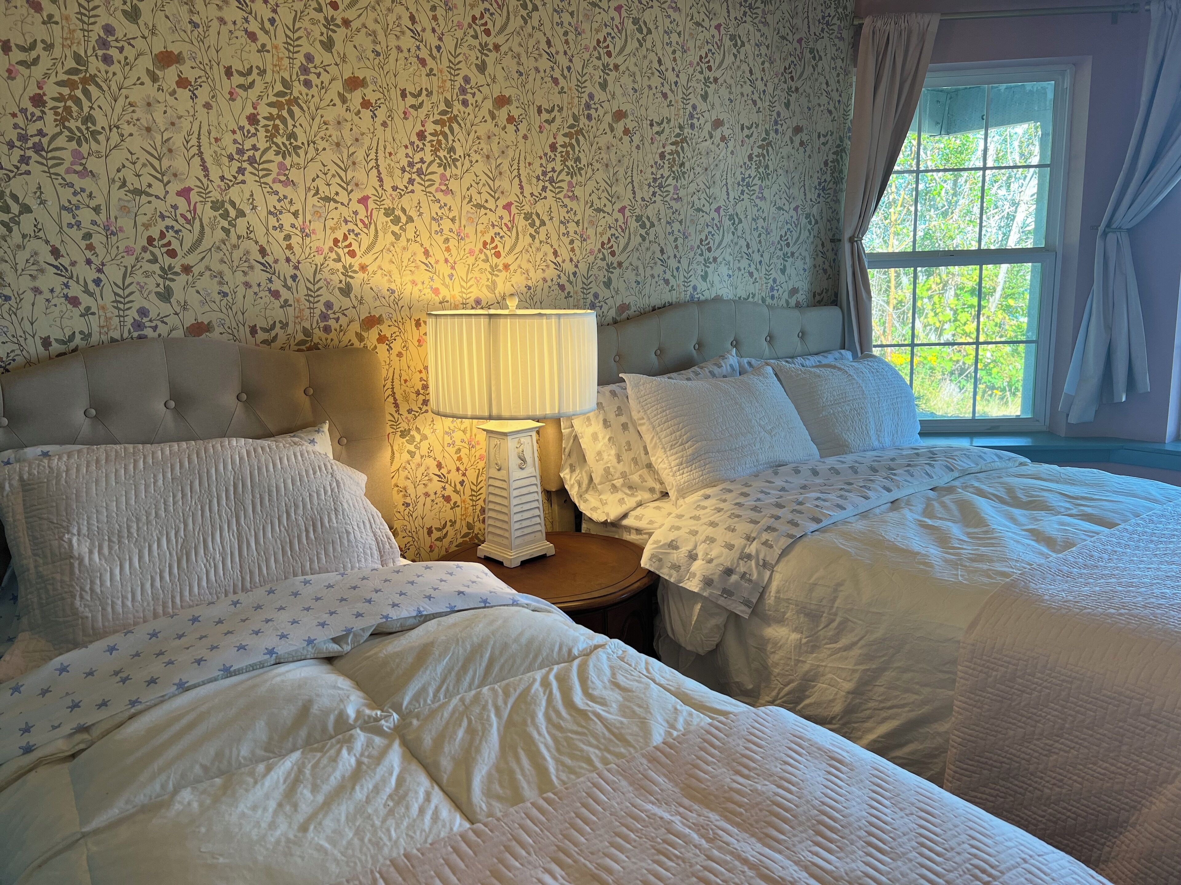 Une chambre avec deux lits, une table de chevet avec une lampe, un papier peint à motifs floraux et une fenêtre avec des rideaux.