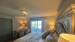 A bedroom with a large bed, a desk, and a chair, a chandelier, and a view through a window.