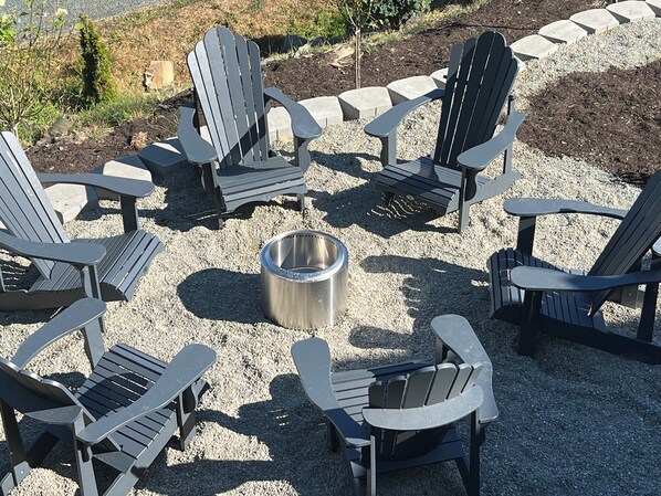 A circular arrangement of Adirondack chairs around a metal fire pit in a gravel area.