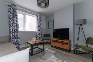 Una sala de estar moderna con un sofá, una mesa de centro, un televisor sobre un mueble de madera, una lámpara de pie y una ventana con cortinas.