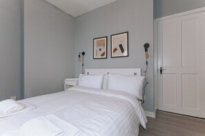 Un dormitorio con cama, almohadas blancas, una mesita de noche, una lámpara de pared y una puerta.