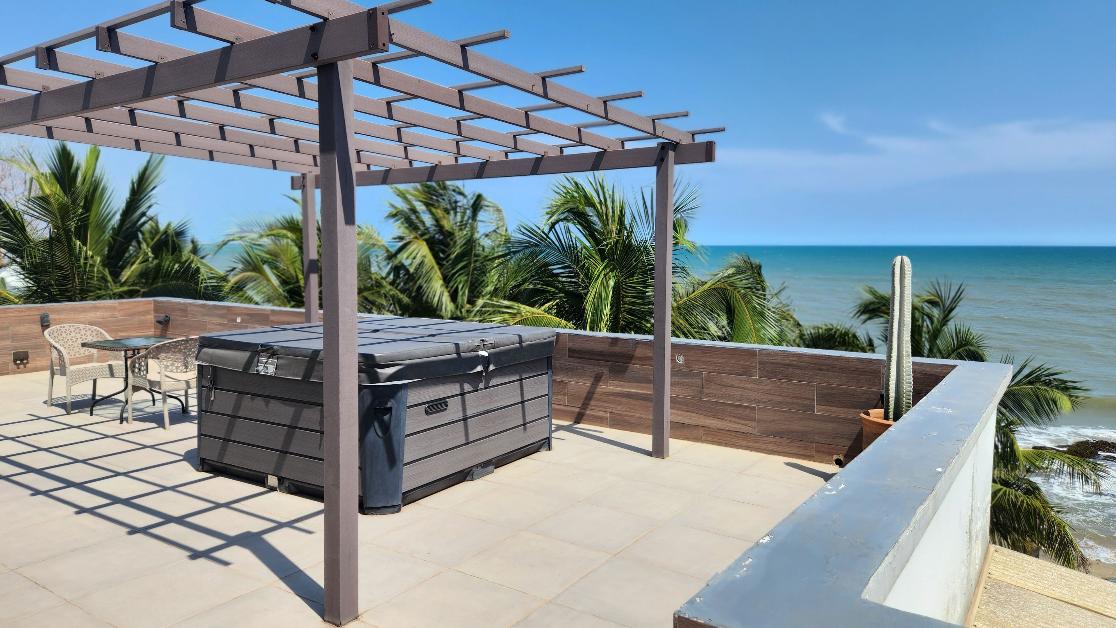Eine Terrasse mit einer Pergola, Sitzgelegenheiten im Freien und einem Whirlpool mit Blick auf einen Strand.
