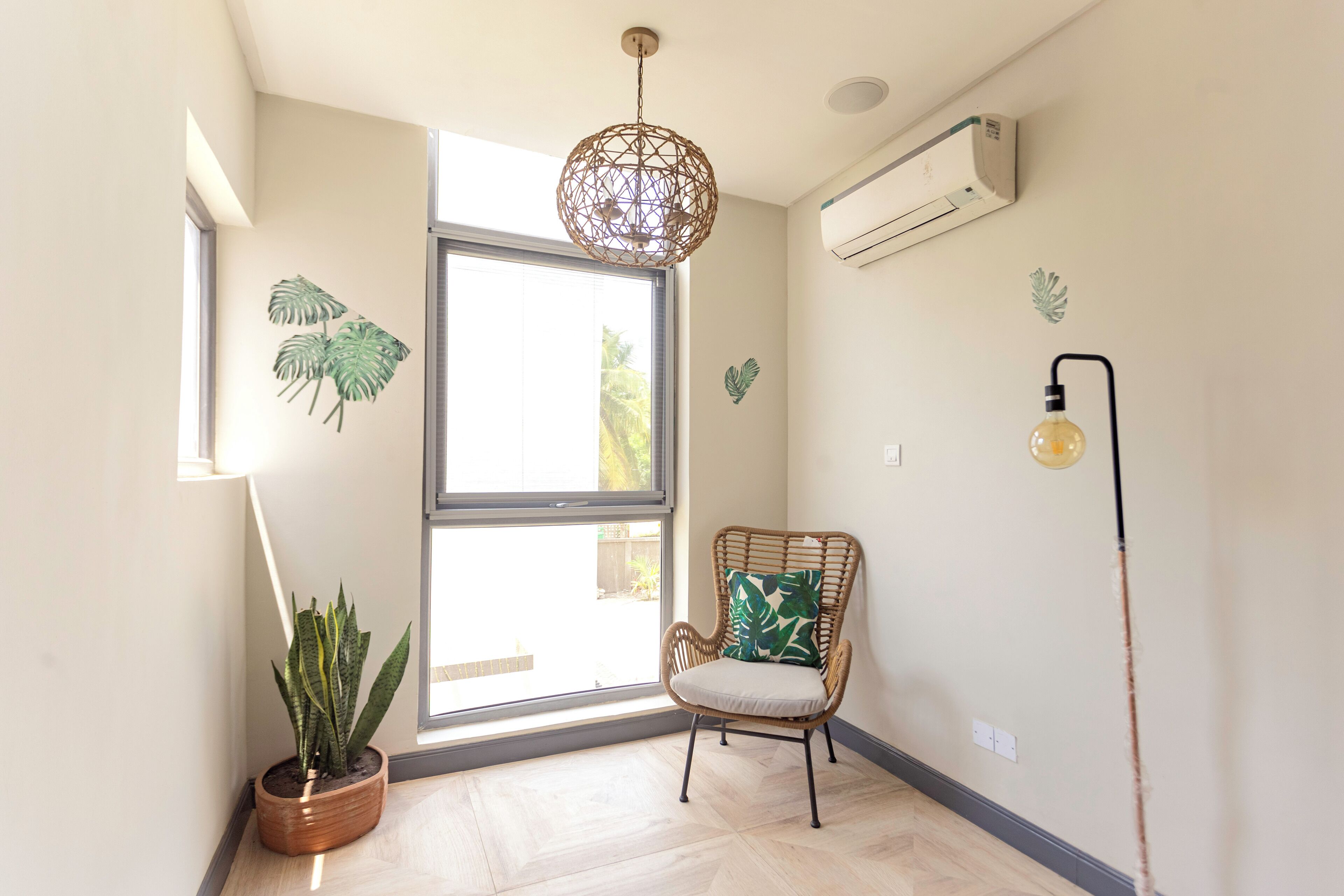 Ein Zimmer mit großem Fenster, einem Korbsessel mit Kissen, einer Topfpflanze und einer Wand-Klimaanlage.