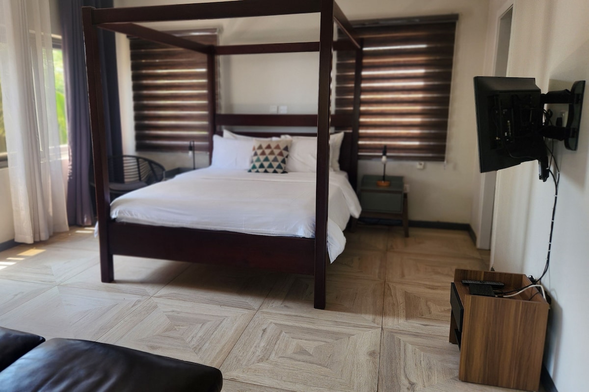 A bedroom with a four-poster bed, a television mounted on the wall, and a view of the outdoors through the window.