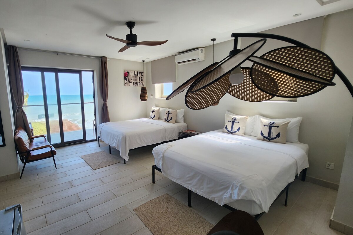 A hotel room with two beds, a ceiling fan, a large window with a view of the beach, and a desk with a chair.