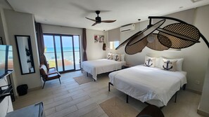 A hotel room with two beds, a ceiling fan, a large window with a view of the beach, and a desk with a chair. - Serenity Residences & Spa - Kokrobite (Kokrobite)