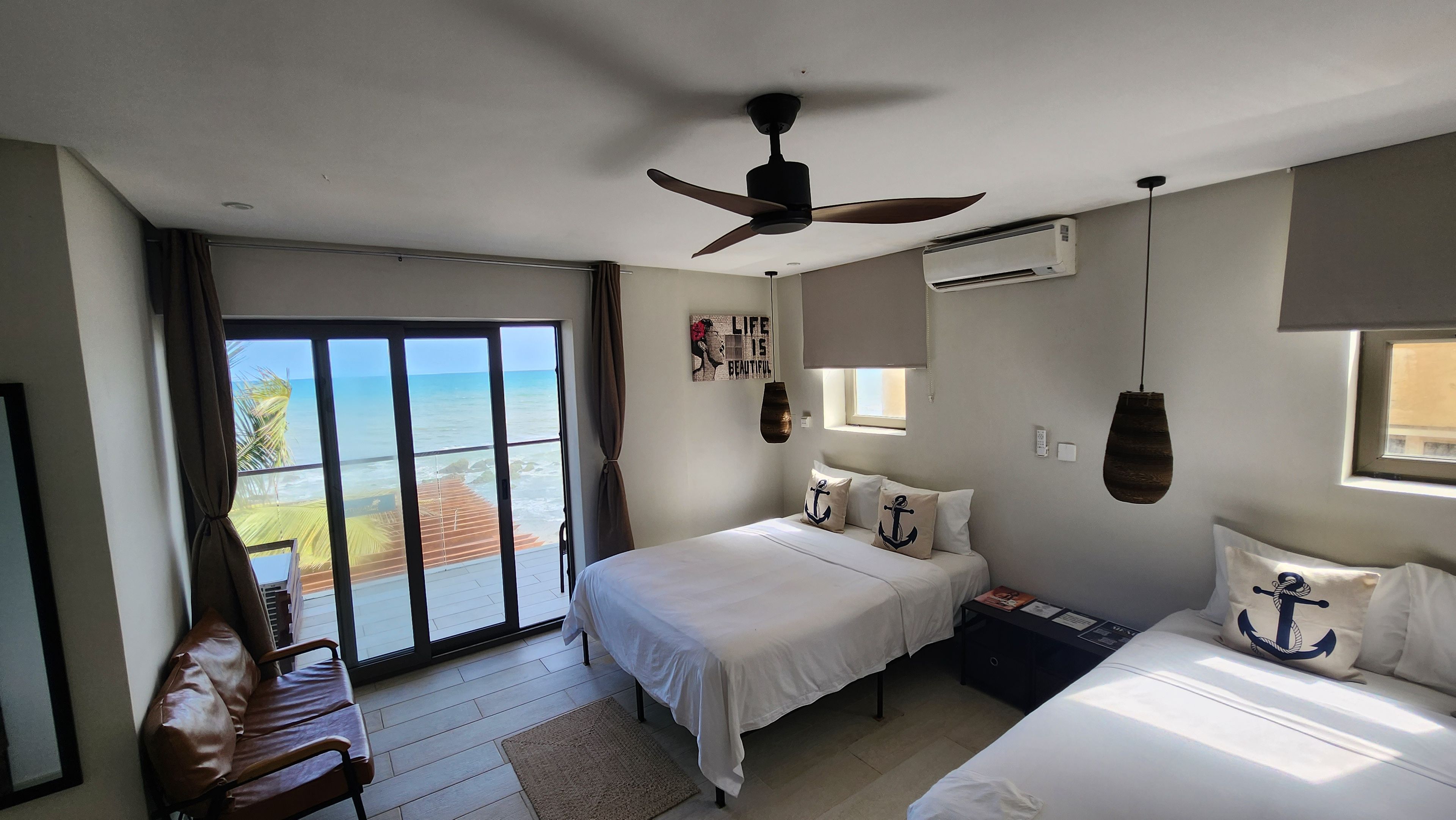 A hotel room with a bed, a desk, a ceiling fan, an air conditioning unit, and a view of the sea through a sliding door.