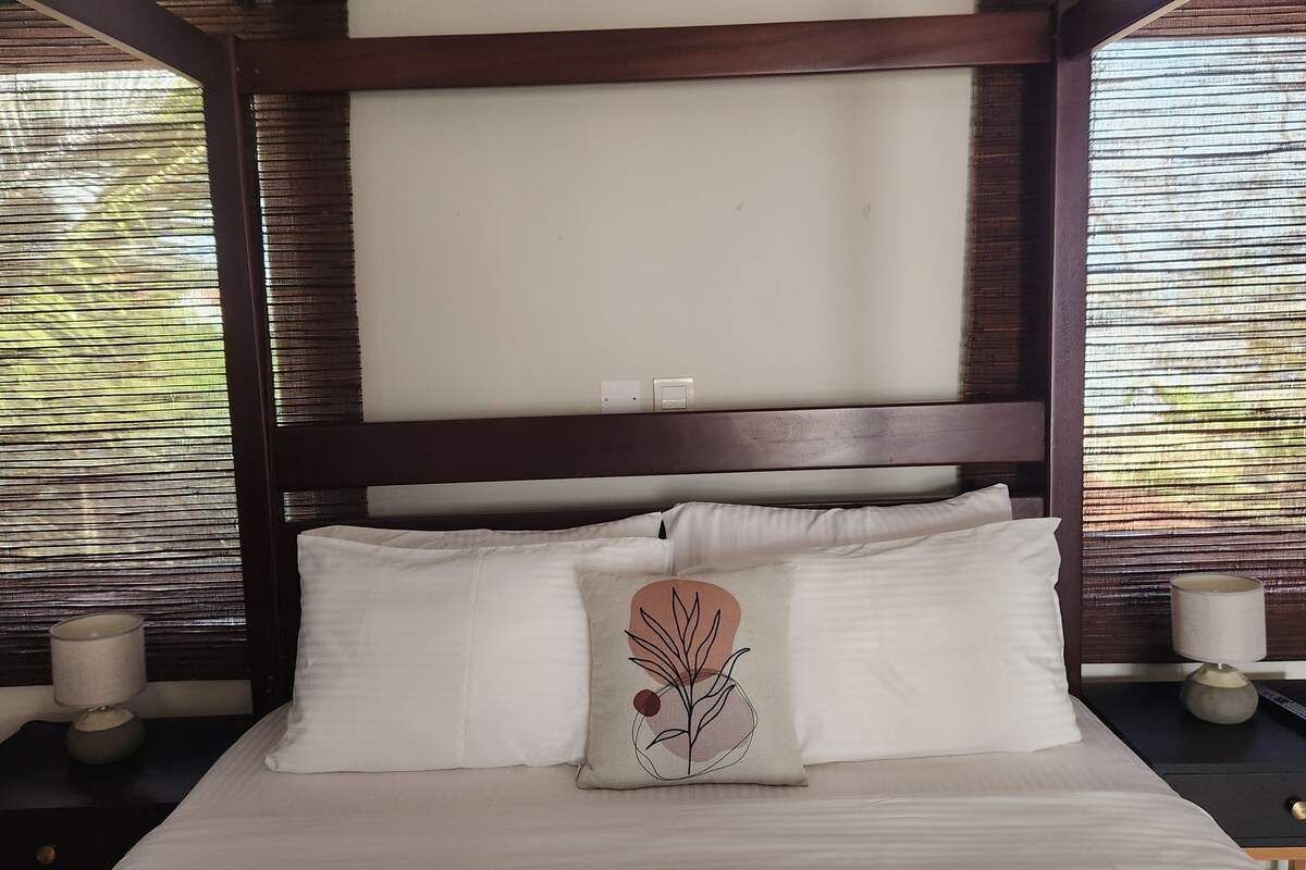 A bed with white bedding, a headboard with a dark finish, and a decorative pillow with a plant design.