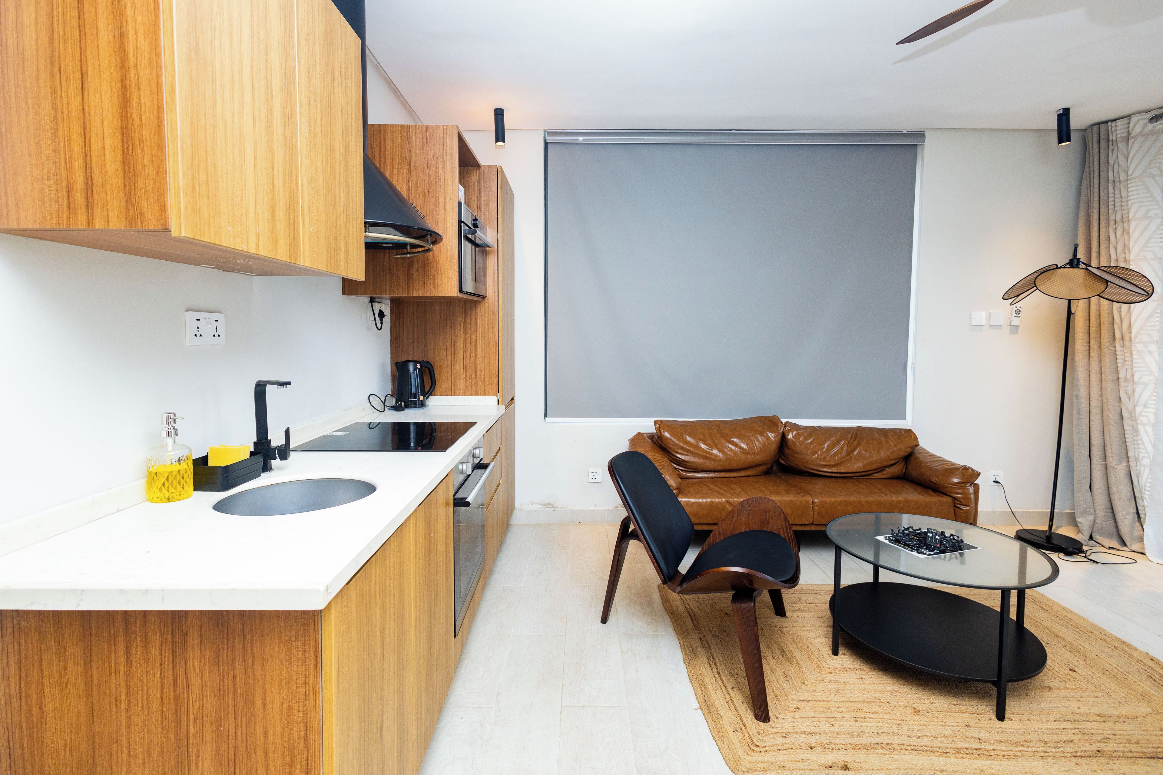 A compact kitchen with a sink, a coffee maker, and wooden cabinets. There is a brown leather sofa and a small round table with a lamp.