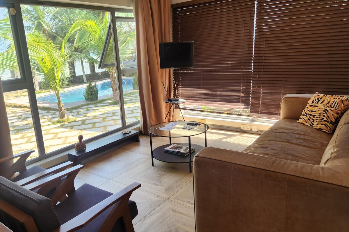 Ein modernes Wohnzimmer mit einer Couch, einem Couchtisch und einem Fernseher. Ein großes Fenster mit Vorhängen bietet Blick auf einen Pool und Palmen.